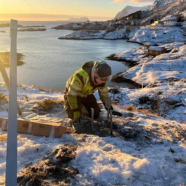 Person i arbeidsklær jobber med betongfester i snødekt terreng med fjordutsikt i solnedgang.