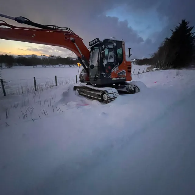 Oransje gravemaskin står i dyp snø ved åkerkant i skumringslys.