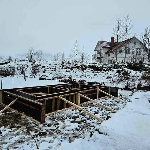 Snødekt byggeplass med reisverk i tre foran hus og gravemaskin i bakgrunnen.