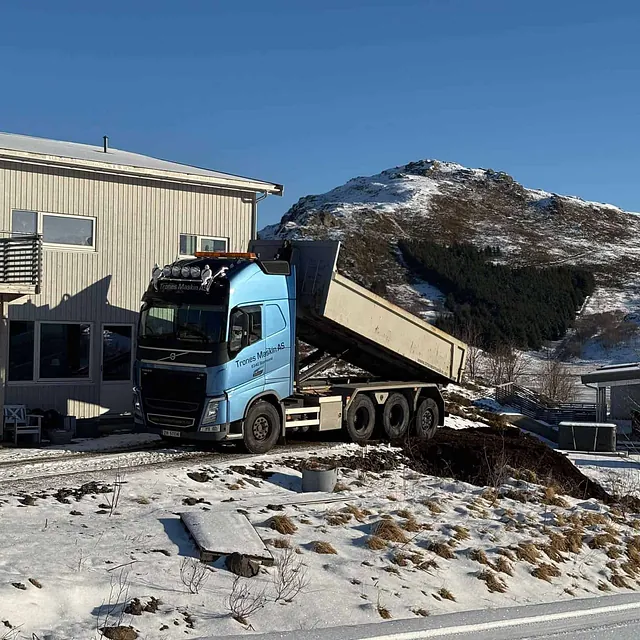 Lastebil med blå førerhytte tømmer masse foran hus i vinterlandskap.