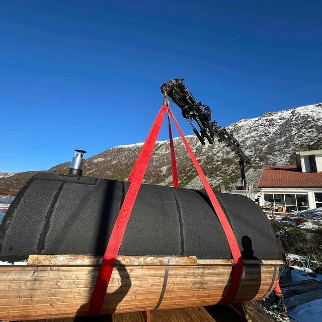 Tønneformet badstue løftes med kran foran snødekte fjell og blå himmel.