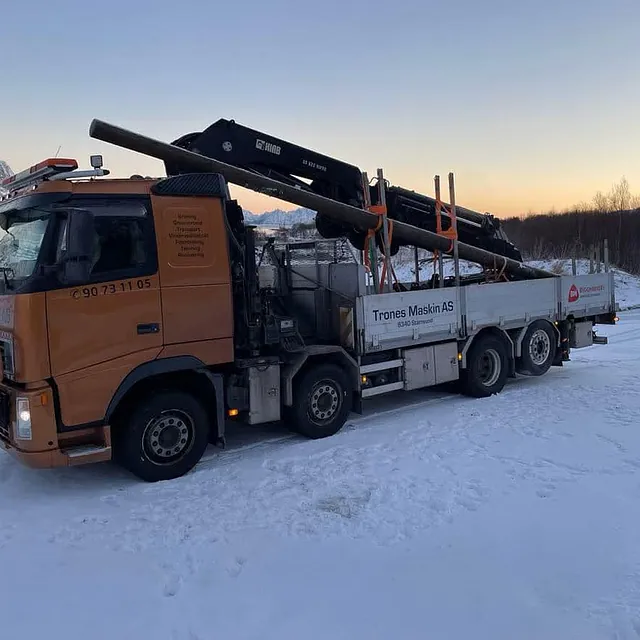 Lastebil fra Trones Maskin med kran og rør lastet bakpå står parkert i snødekt landskap.