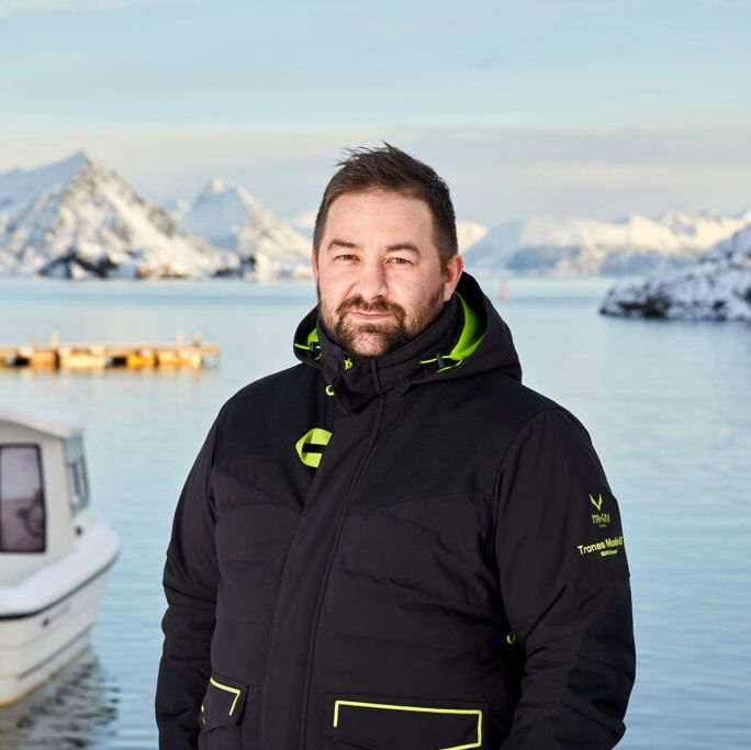 Thomas Andre Trones i svart jakke står ved stille vinterfjord omgitt av båter og snødekte fjell.